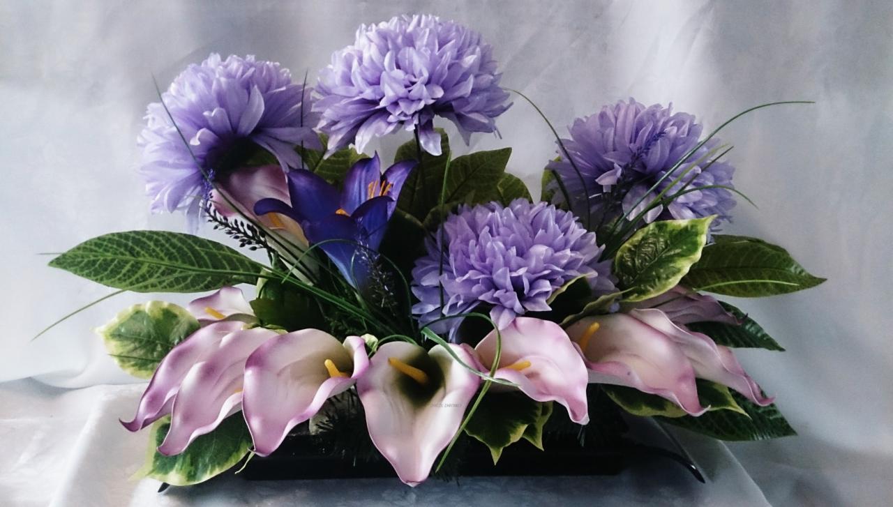 Kompozycja kwiatowa z lilii calla i kulistych kwiatów w odcieniach fioletu, umieszczona w czarnym, niskim wazonie na tle białej tkaniny.