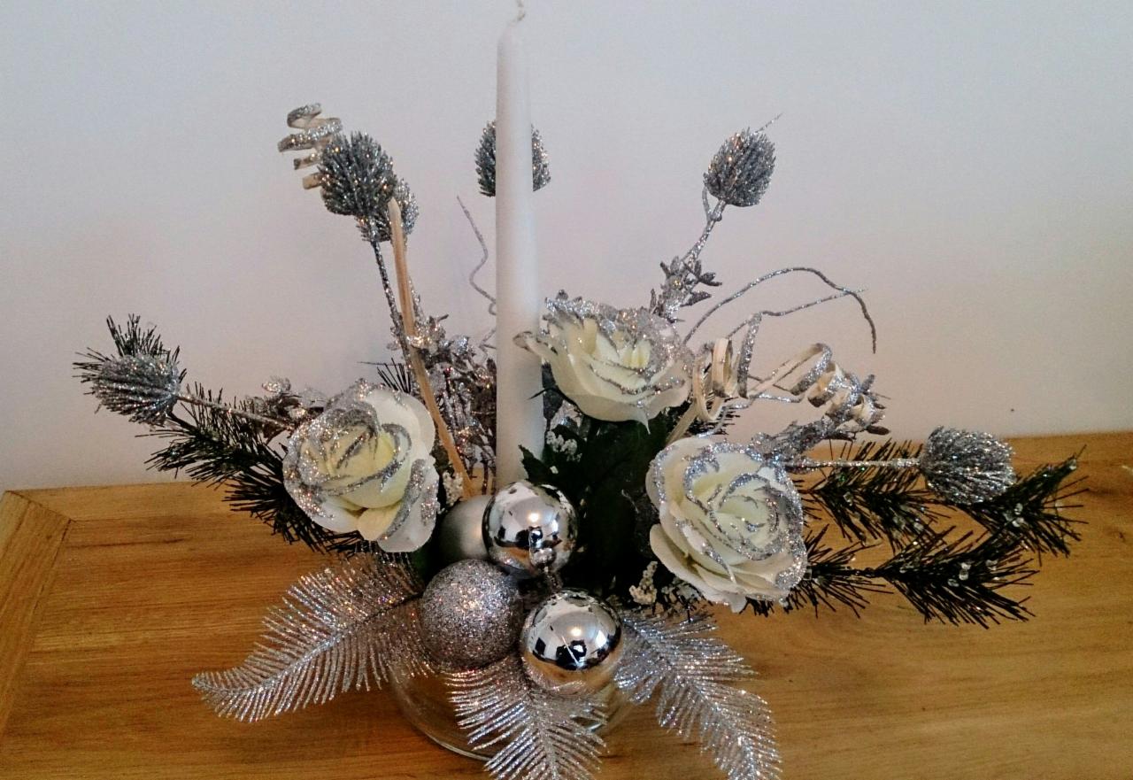 Świąteczna kompozycja ze srebrnymi ozdobami, białymi różami i świecą, wykonana w Radzyniu Podlaskim, umieszczona na drewnianym stole.
