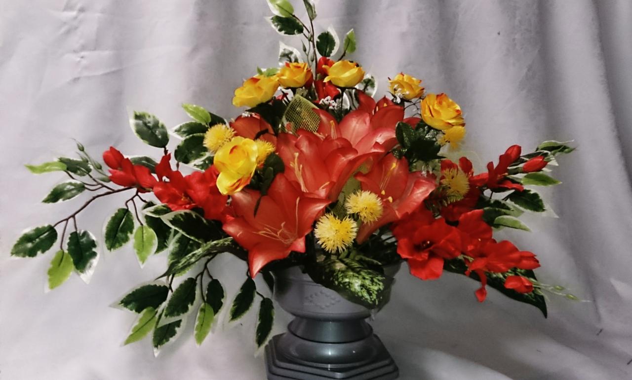 Dekoracyjny bukiet sztucznych kwiatów w odcieniach czerwieni i żółci, umieszczony w szarej, plastikowej doniczce na tle białej tkaniny.