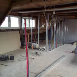 Poddasze w trakcie remontu: metalowa konstrukcja ścianki działowej, płyta karton gips oparta o ścianę, przewody instalacyjne w ścianie, widoczne okna dachowe i narzędzia pracy.