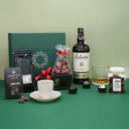 Elegancki zielony kosz prezentowy z whisky Ballantine's 21, kawą, czekoladkami, figami w brandy oraz filiżanką kawy na zielonym tle.