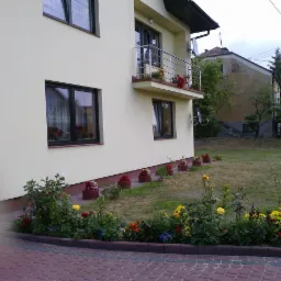 Żółta elewacja domu z ciemnymi oknami i balkonem z kwiatami, numer domu '7', rabata kwiatowa wzdłuż ścieżki z kostki brukowej, trawnik i budynek w tle.