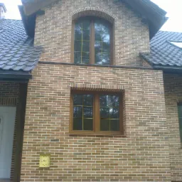 Elewacja domu z brązowej cegły klinkierowej, z brązowymi oknami i drzwiami. Widoczne okna dachowe i komin. Detal architektoniczny w stylu rustykalnym.