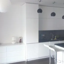 Nowoczesna, minimalistyczna kuchnia z białymi frontami i czarnym blatem, oświetlona wiszącymi lampami kulistymi. Widoczny biały kredens i ozdobne figurki.