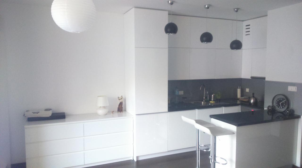 Nowoczesna, minimalistyczna kuchnia z białymi frontami i czarnym blatem, oświetlona wiszącymi lampami kulistymi. Widoczny biały kredens i ozdobne figurki.