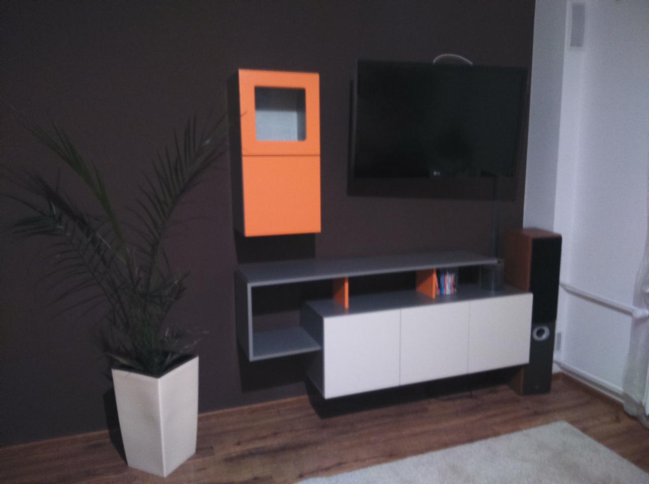 Nowoczesna aranżacja salonu z wiszącą szafką RTV, pomarańczową szafką i telewizorem na brązowej ścianie. Uzupełnieniem jest roślina w białej donicy.