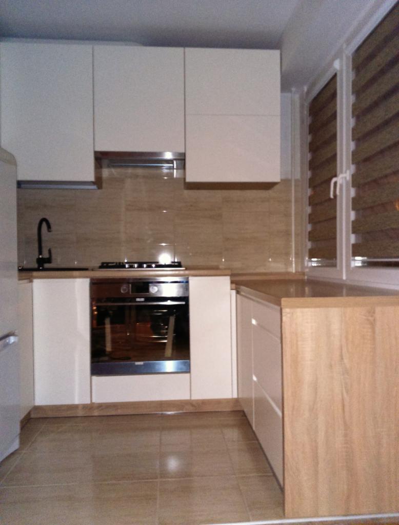 Jasna kuchnia z białymi szafkami i drewnianym blatem, z piekarnikiem w zabudowie. Widoczne rolety w oknie i beżowe płytki na ścianie. Ujęcie z niskiego kąta.