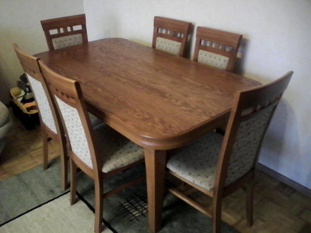 Rozkładany stół z drewna i cztery krzesła z tapicerowanymi oparciami. Widoczny fragment dywanu i ściany w tle. Oświetlenie naturalne.