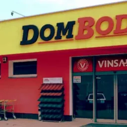 Fasada budynku DOMBOD bis z żółto-czerwonymi ścianami. Sklep budowlany VINSAR z wózkami i ekspozycją dachówek przed wejściem.