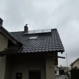 Dom jednorodzinny z ciemną dachówką, oknem dachowym i panelami solarnymi na dachu. Widok z dołu na fragment elewacji i dach w pochmurny dzień.