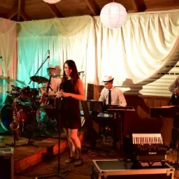 Zespół muzyczny w białych kapeluszach na scenie, wokalistka w czarnej sukience, saksofonista, perkusista, klawiszowiec i gitarzysta, białe zasłony w tle, oświetlenie sceniczne.