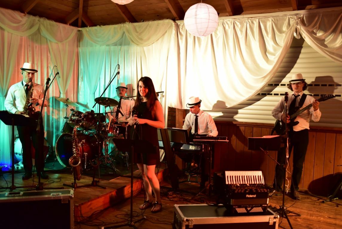 Zespół muzyczny w białych kapeluszach na scenie, wokalistka w czarnej sukience, saksofonista, perkusista, klawiszowiec i gitarzysta, białe zasłony w tle, oświetlenie sceniczne.