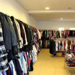 Wnętrze sklepu z odzieżą używaną. Rzędy ubrań wiszą na wieszakach wzdłuż ścian. Widoczne kurtki, koszule i sukienki. Jasne oświetlenie, beżowa podłoga z płytek.