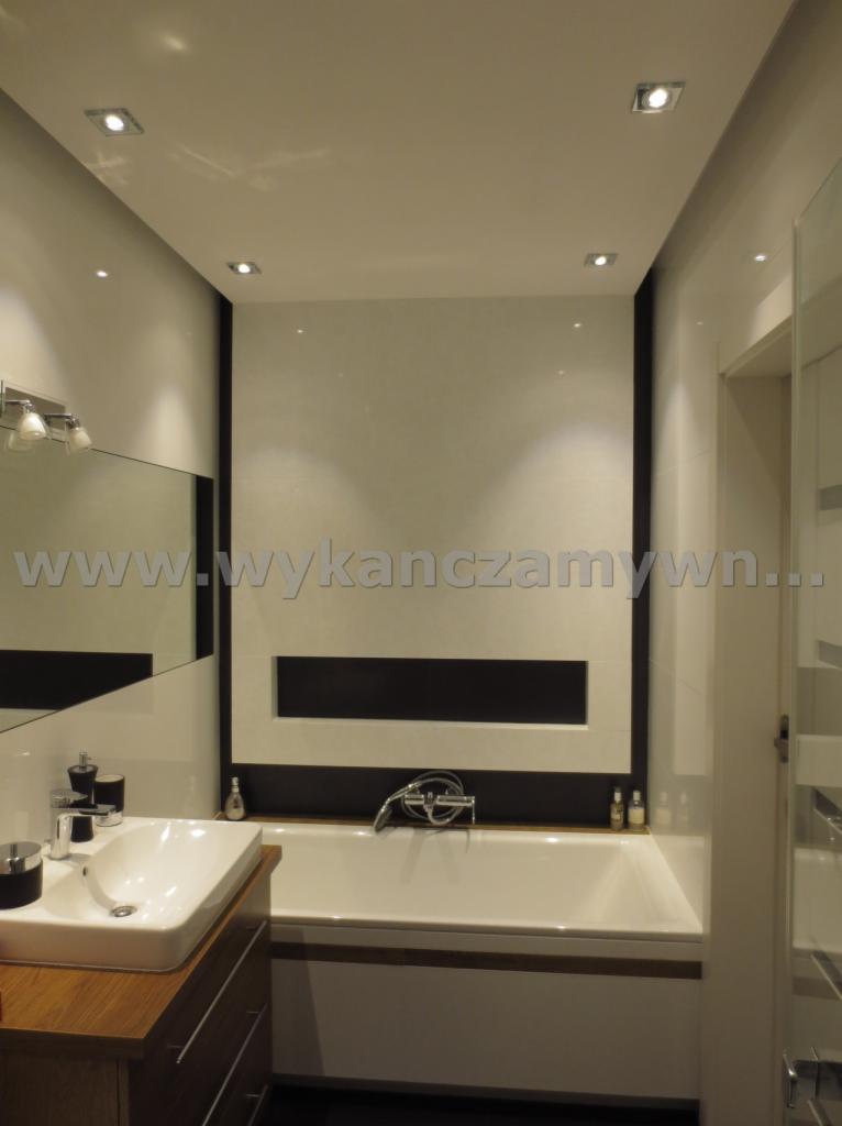 Nowoczesna łazienka z wanną w zabudowie, umywalką na drewnianej szafce i lustrem. Białe płytki z czarnymi akcentami i oświetleniem punktowym.