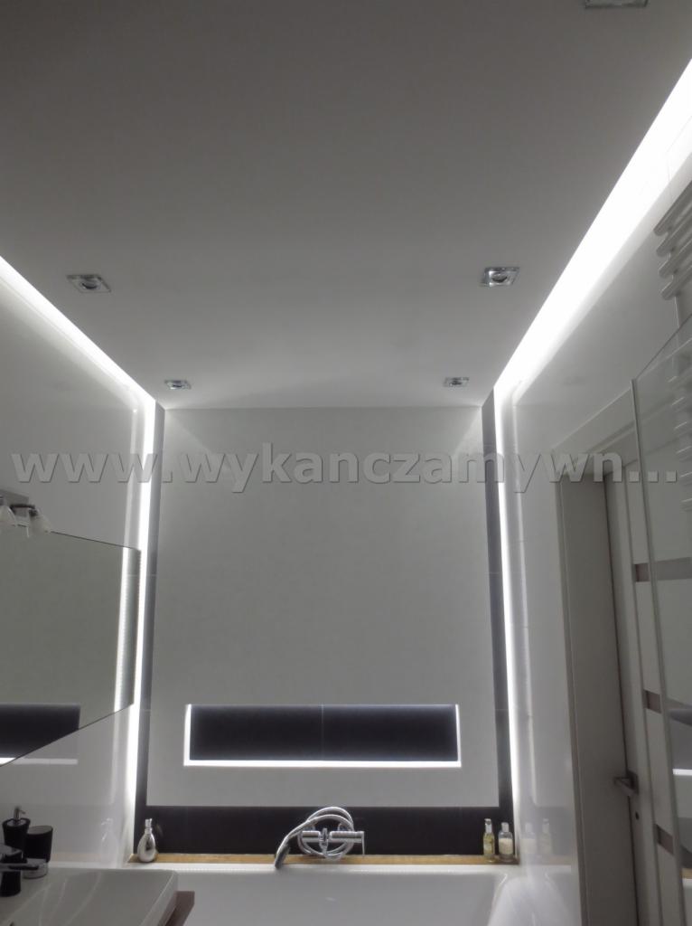 Nowoczesna łazienka z białymi ścianami, podświetlanym sufitem i roletą w oknie. Widoczna armatura i lustro. Minimalistyczny design z dbałością o detale.