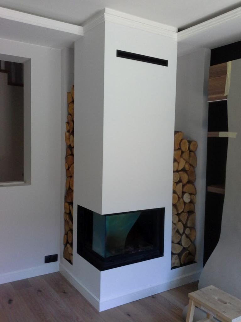 Nowoczesny kominek narożny z przeszklonym paleniskiem, obudowany na biało, z dekoracyjnym układem drewna po bokach. Minimalistyczny design, jasne wnętrze.