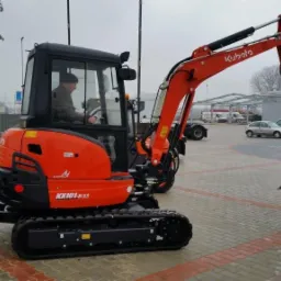 Pomarańczowa minikoparka Kubota KX101-3a3 na gąsienicach z operatorem w kabinie, zaparkowana na kostce brukowej, na tle parkingu i zabudowań.