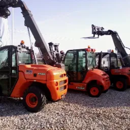 Pomarańczowe ładowarki teleskopowe AUSA na żwirowym podłożu, z podniesionymi ramionami i widłami. Częściowo widoczny biały van w tle.