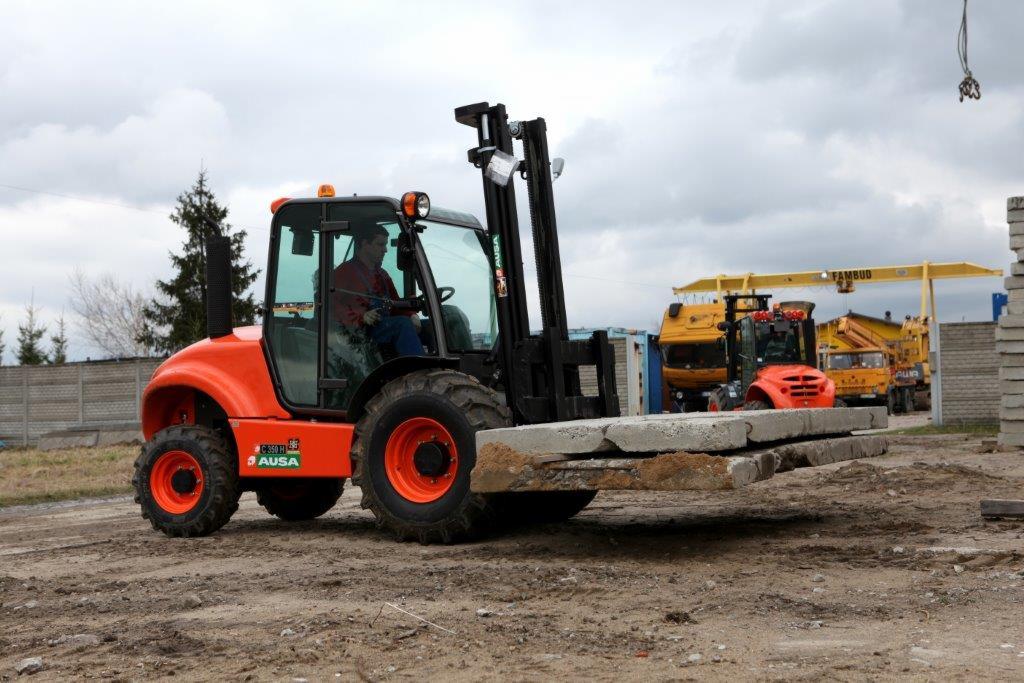 Pomarańczowa ładowarka teleskopowa AUSA z operatorem, przewożąca betonowe płyty na placu budowy. W tle inne maszyny i żuraw.