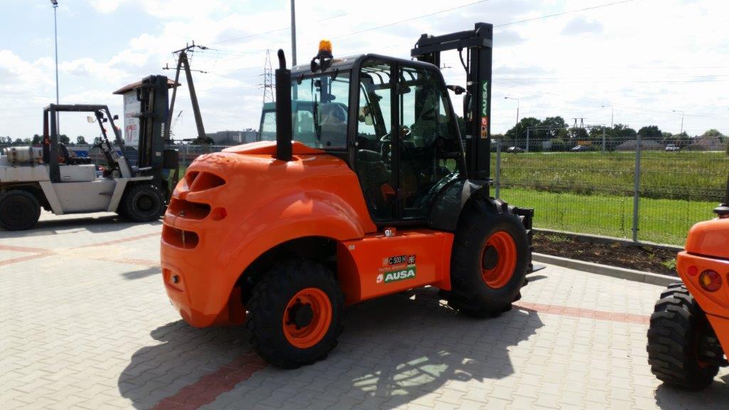 Pomarańczowy wózek widłowy AUSA C 500 H na kostce brukowej, z widocznymi innymi wózkami w tle i zielenią. Charakterystyczny design, z kabiną i podnośnikiem.