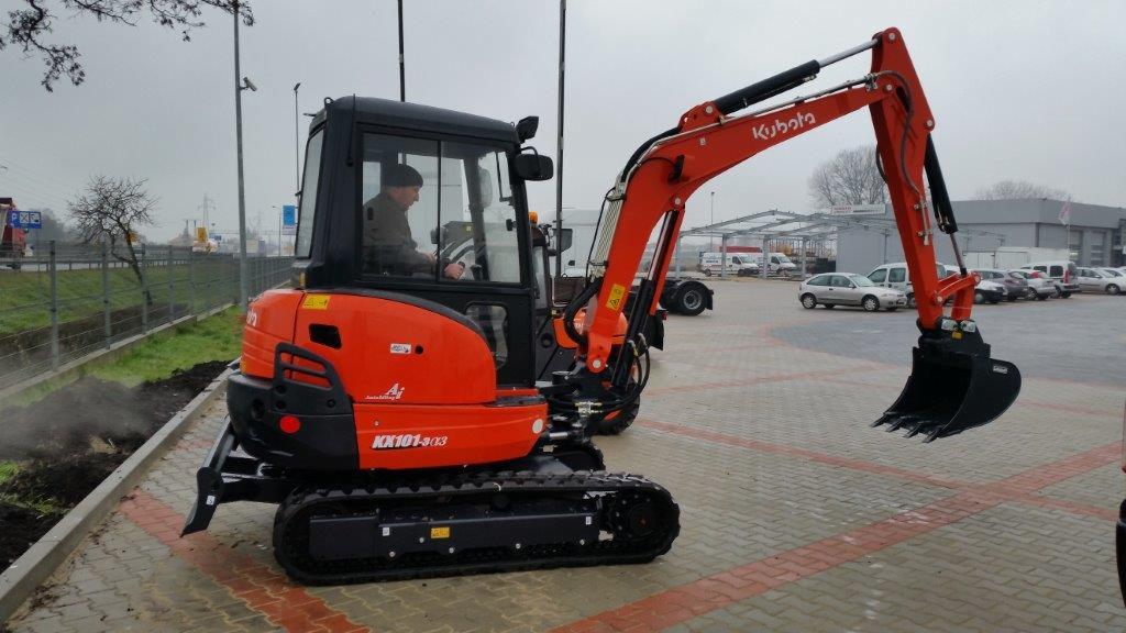 Pomarańczowa minikoparka Kubota KX101-3a3 na gąsienicach z operatorem w kabinie, zaparkowana na kostce brukowej, na tle parkingu i zabudowań.