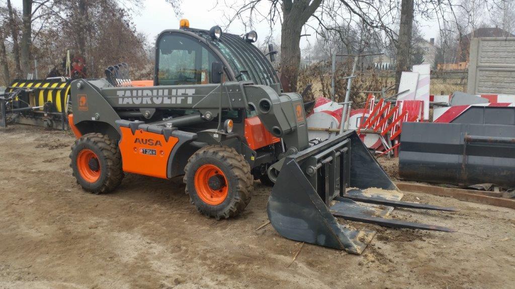 Szaro-pomarańczowa ładowarka teleskopowa AUSA Taurulift T204H z łyżką i widłami na nierównym terenie, jesienne drzewa w tle. Sprzęt budowlany.