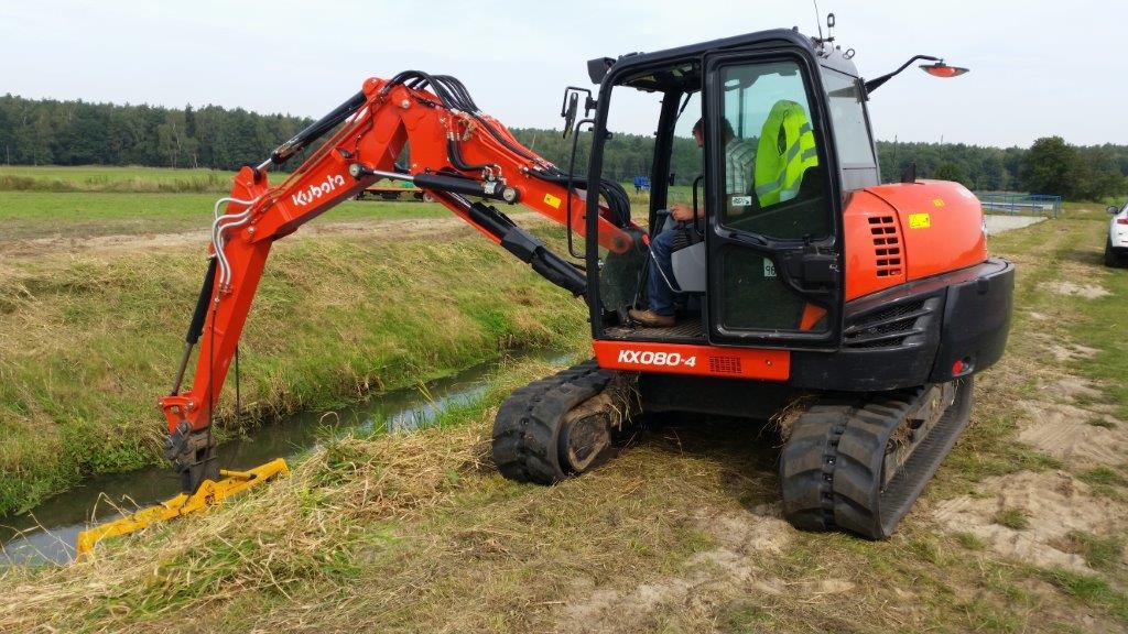 Pomarańczowo-czarna minikoparka Kubota KX080-4 z operatorem w kabinie, w trakcie prac ziemnych przy rowie melioracyjnym na tle zieleni i drzew.