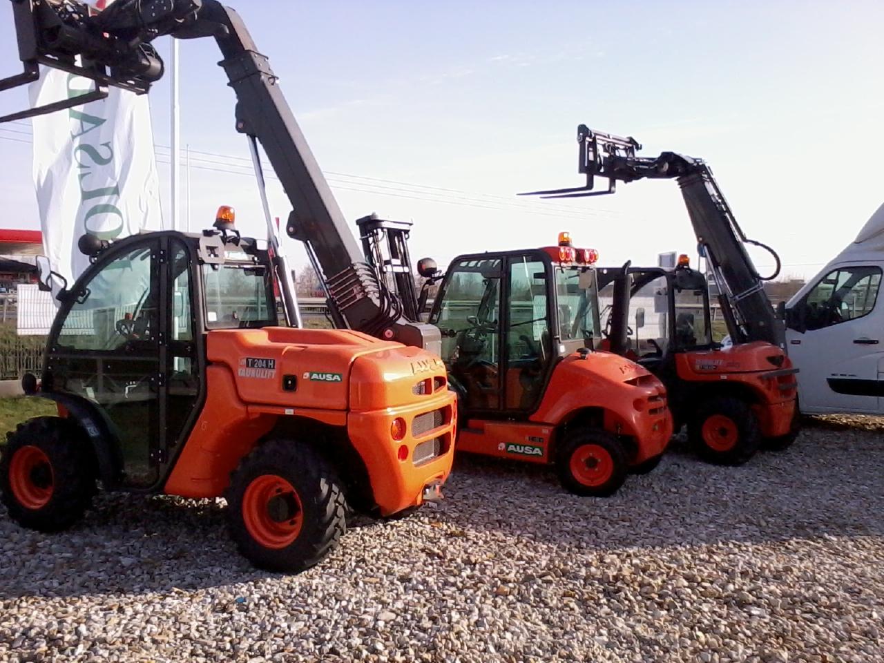 Pomarańczowe ładowarki teleskopowe AUSA na żwirowym podłożu, z podniesionymi ramionami i widłami. Częściowo widoczny biały van w tle.