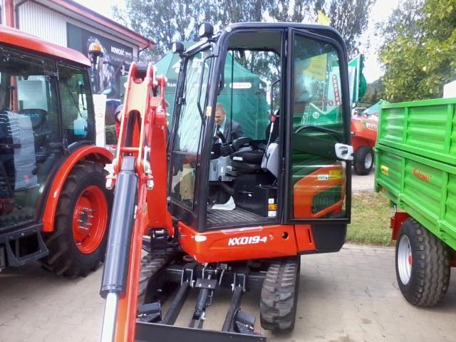 Czerwono-czarna minikoparka KX019-4 z kabiną, widoczna na tle ekspozycji targowej z innymi maszynami rolniczymi i traktorem. Widok z boku, na pierwszym planie ramię koparki.