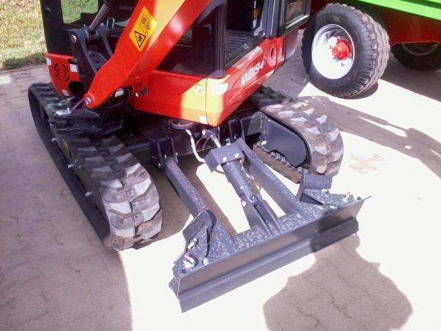 Czerwona minikoparka gąsienicowa z przodu, z czarnym lemieszem, częściowo widoczna przyczepa z kołem w tle. Maszyna budowlana na betonowej powierzchni.