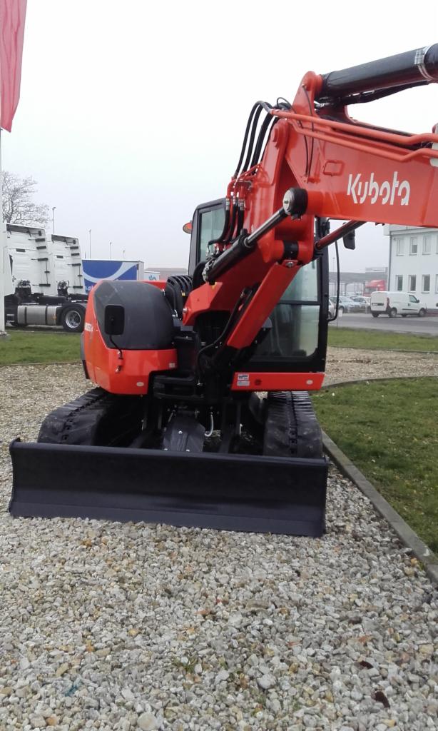 Pomarańczowa minikoparka Kubota z czarnym lemieszem na żwirowym podłożu. Widoczne detale hydrauliki i gąsienic. W tle budynki i zaparkowane pojazdy.