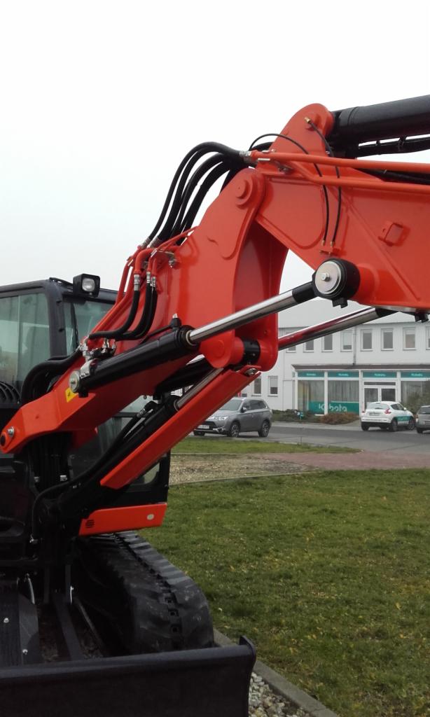 Pomarańczowa minikoparka Kubota z widocznym ramieniem, siłownikami hydraulicznymi i gąsienicami. W tle budynek z logo Kubota i zaparkowane samochody.