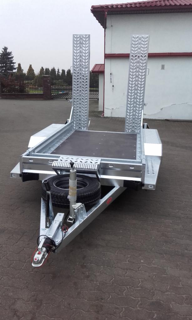 Nowa, srebrna przyczepa niskopodwoziowa z najazdami, zaparkowana na zewnątrz. Widoczny zapasowy koło i zaczep. Tło stanowi budynek i zieleń.