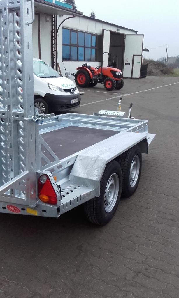 Przyczepa transportowa z rampą najazdową, widoczna z tyłu, z pomarańczowym traktorem i białym samochodem w tle na zewnątrz budynku.
