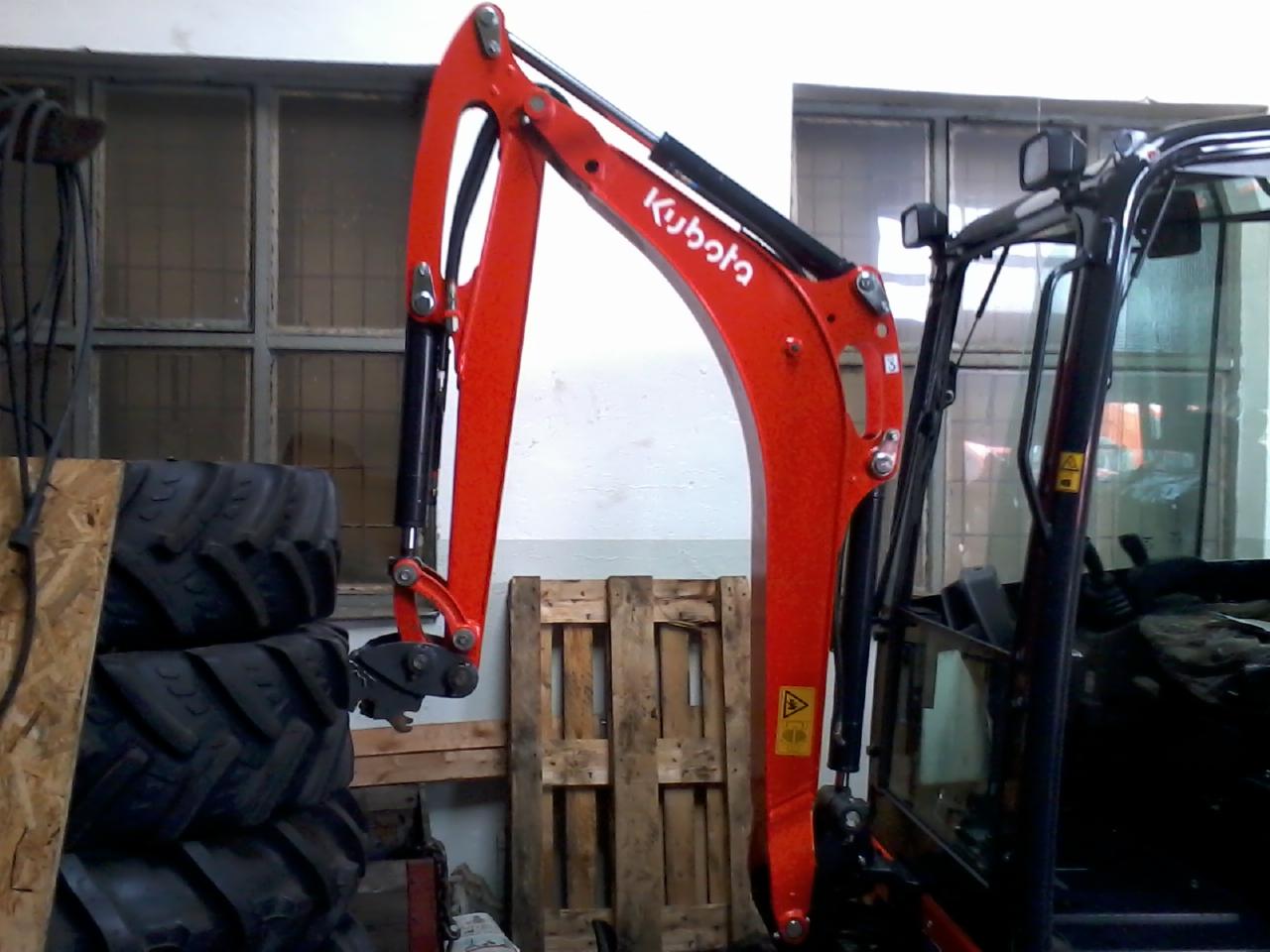 Częściowo widoczna kabina i ramię koparki Kubota w kolorze czerwonym, na tle okna i sterty opon. Widoczna paleta. Ujęcie wewnątrz pomieszczenia.