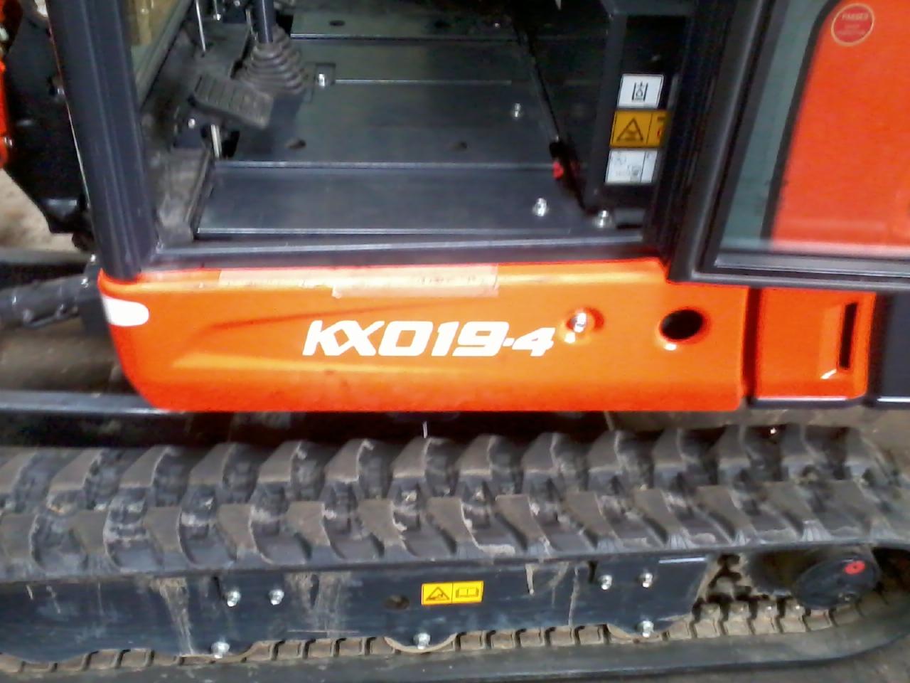 Pomarańczowa minikoparka Kubota KX019-4 z widocznymi gąsienicami, kabiną i oznaczeniami bezpieczeństwa. Detal z bliska, częściowy widok maszyny.