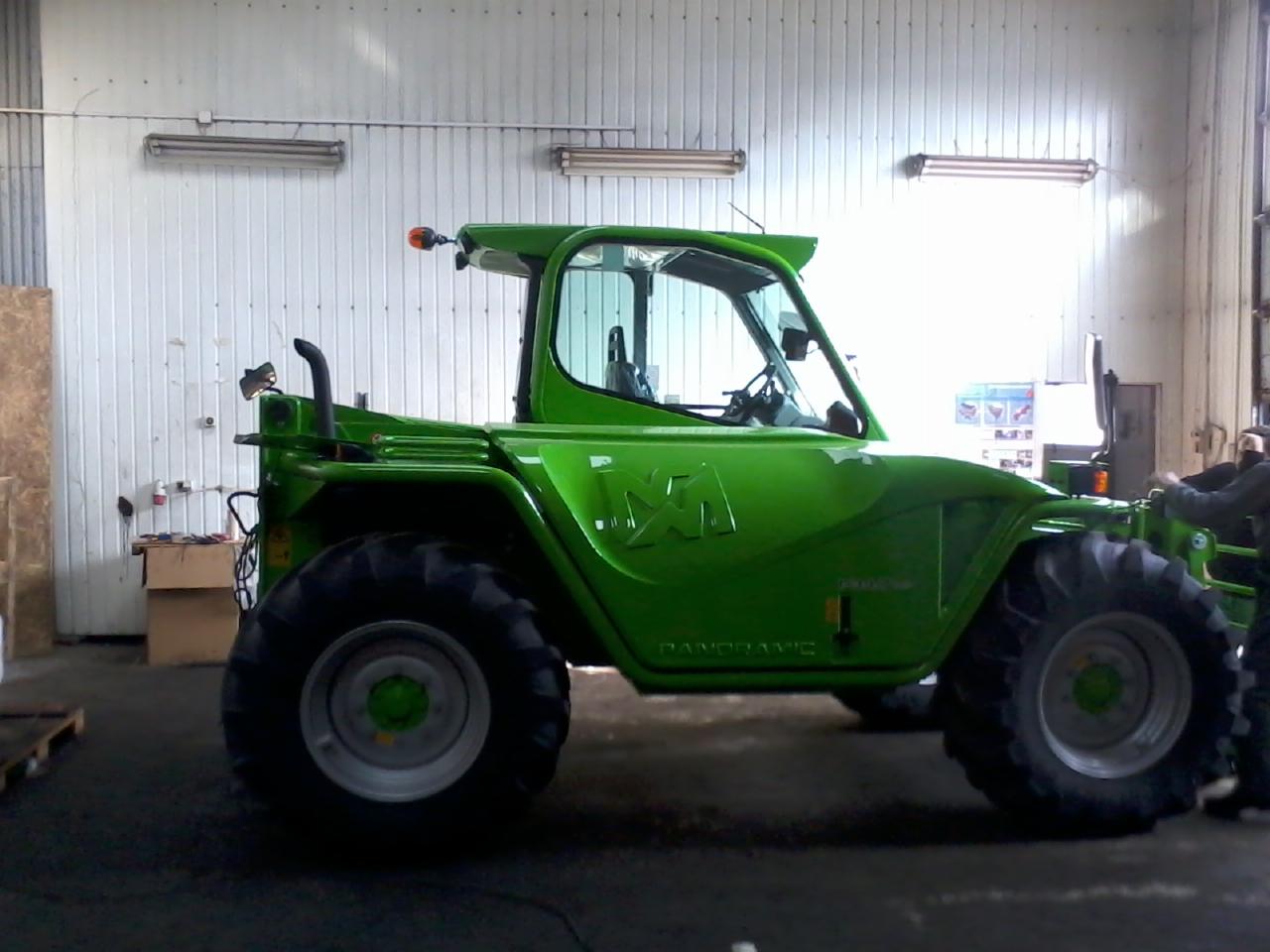 Zielona ładowarka teleskopowa Merlo Panoramic z dużymi oponami, widok z boku w hali z blachy falistej. Widoczny fragment osoby, skrzynki i oświetlenie.