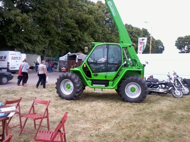 Zielona ładowarka teleskopowa Merlo Turbo Farmer na trawie, obok zaparkowane motocykle, w tle osoby i budynki, dzień.