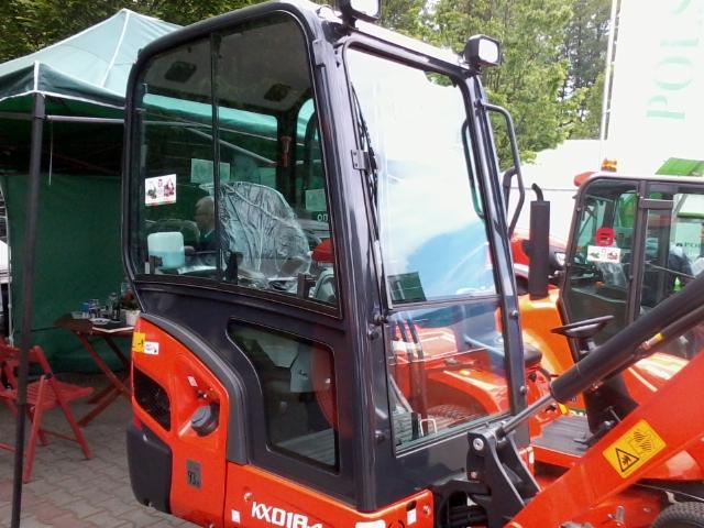 Pomarańczowa minikoparka Kubota KX018-4 z kabiną, widoczna z bliska, na tle zielonego namiotu i innych maszyn. Kabina oklejona naklejkami informacyjnymi.