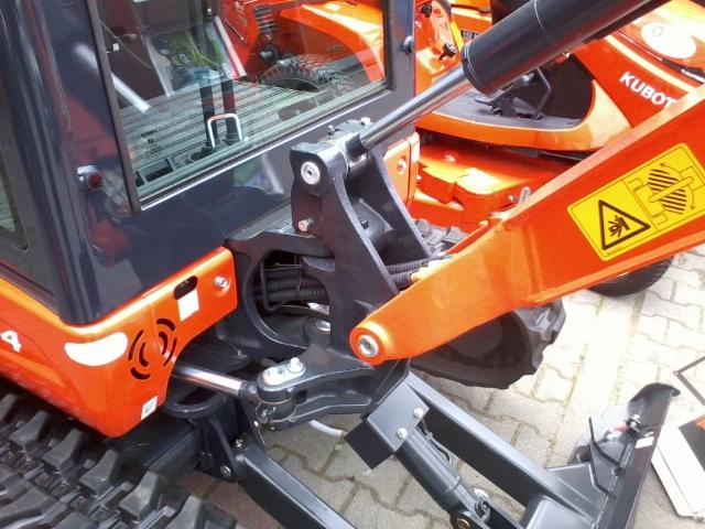 Pomarańczowa minikoparka Kubota z czarnymi elementami, widoczne detale konstrukcyjne i ostrzegawcza naklejka. Ujęcie z bliska, częściowo widoczna inna maszyna w tle.