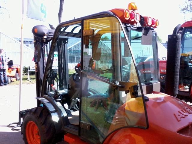 Pomarańczowa ładowarka teleskopowa z kabiną, widoczne detale konstrukcyjne i oświetlenie, tło z zielenią i fragmentem szklarni. Prezentacja detali urządzenia.