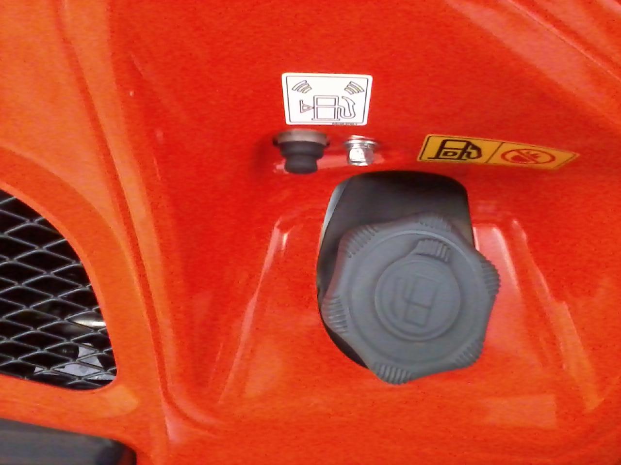 Pomarańczowa obudowa maszyny budowlanej z czarnym wlewem paliwa i naklejkami informacyjnymi. Widoczny fragment siatki wentylacyjnej.
