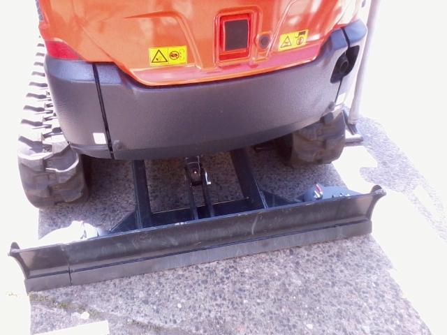 Pomarańczowa minikoparka widziana z tyłu, z widocznymi gąsienicami i lemieszem, stojąca na szarym betonowym podłożu w jasnym świetle.