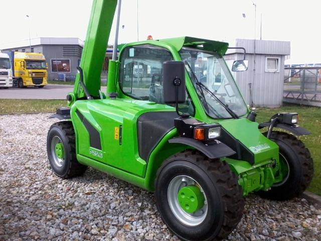 Jasnozielona ładowarka teleskopowa Merlo P25.6 na żwirowym podłożu. Widoczny fragment podniesionego ramienia. W tle budynek i żółta ciężarówka.