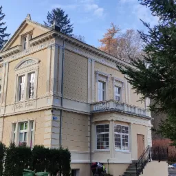 Odnowiona, beżowa kamienica z dekoracyjnymi elementami architektonicznymi, widoczna spod drzew, z fragmentem prac porządkowych na pierwszym planie.