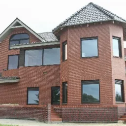 Dom z czerwonej cegły z czarnymi ramami okiennymi i częściowo opuszczonymi roletami zewnętrznymi, widok z zewnątrz na fragment elewacji z oknami o różnych rozmiarach i kształtach.