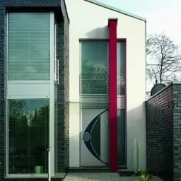 Nowoczesny dom z dużymi oknami z żaluzjami i drzwiami zewnętrznymi z oryginalnym, półokrągłym wzorem w kolorach granatowym i zielonym, podkreślonym pionowym, czerwonym elementem architektonicznym.