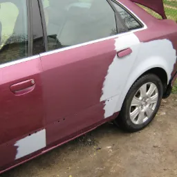 Bordowy samochód osobowy w trakcie naprawy karoserii, z widocznymi obszarami pokrytymi szpachlą, przygotowany do lakierowania, z otwartą klapą bagażnika i tłem z trawy i siatki ogrodzeniowej.