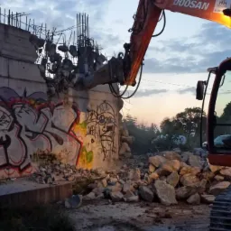 Pomarańczowy koparka Doosan kruszy betonowy mur pokryty graffiti. W tle widoczne niebo o zmierzchu i gruz.