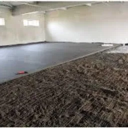 Wnętrze hali w trakcie wylewania posadzki. Część powierzchni pokryta świeżą, szarą wylewką, druga część zbrojona siatką i przygotowana do zalania. Widoczne narzędzia pomiarowe i poziomica.